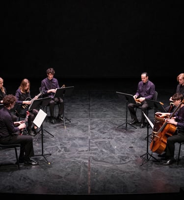 A wide-angle artistic shot of a dark charcoal stage before a performance. Subtle muted plum highlights catch the edges of musical instruments. The atmosphere is expectant and professional.