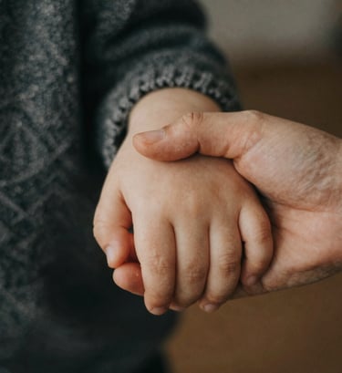 A close-up, candid shot of a young child's hand holding a parent's hand. The lighting is soft and warm, with a shallow depth of field. Soft skin tones against a Charcoal-colored knit sweater. Authentic and intimate.