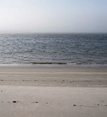 Large vertical landscape photograph of a desolate, mist-covered coastline in Northern Europe. The water is a dark charcoal, the sand a pale stone gray, and the sky a soft white. The composition is extremely minimal, focusing on the horizontal layers of the landscape.