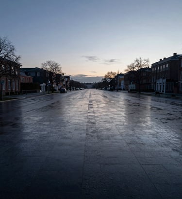 A wide-angle horizontal photograph of an empty city street at dawn. The pavement is a deep charcoal, wet with rain, reflecting the muted slate blue light of the early morning sky. The atmosphere is calm, elegant, and silent.