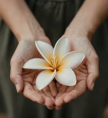 An intimate, artistic macro shot of two hands gently holding a small tropical flower. The focus is soft, highlighting the texture of the petals and the connection between the hands. The lighting is warm and low-contrast, featuring shades of #C7B7A3 and #5F705B.