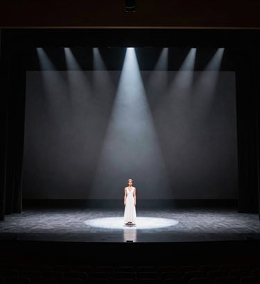 A wide-angle professional photograph of a grand North American / US theater stage during a performance. The composition is minimalist and elegant, featuring a single performer under a crisp spotlight with a deep gray and white color palette.