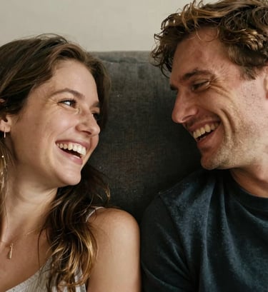 A close-up candid photo of a couple laughing together on a soft charcoal-colored sofa. The lighting is golden and warm, capturing the fine details of their expressions and the empathetic mood of the moment.