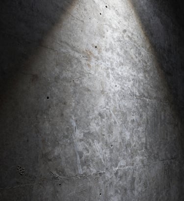An artistic close-up of a textured concrete wall. The surface is a mix of muted slate and deep charcoal tones, with a single beam of pale ice white light grazing the surface to reveal the intricate, porous texture of the stone.