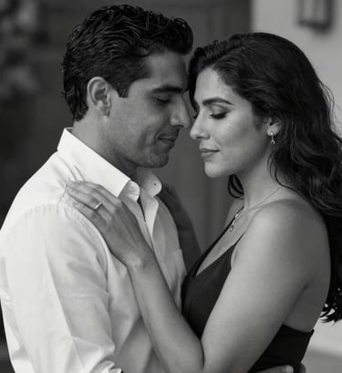 An intimate black and white portrait of a Hispanic / Spanish-speaking couple embracing, with high contrast and soft lighting that highlights the elegance of their attire. The composition is cinematic and romantic.