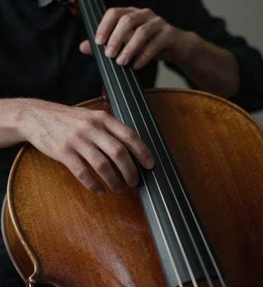Introspective close-up photograph of a person's hands playing a dark wooden cello. The lighting is moody and focused with high contrast, highlighting the texture of the wood and the movement of the fingers. International / Western style, dark brown and charcoal tones.