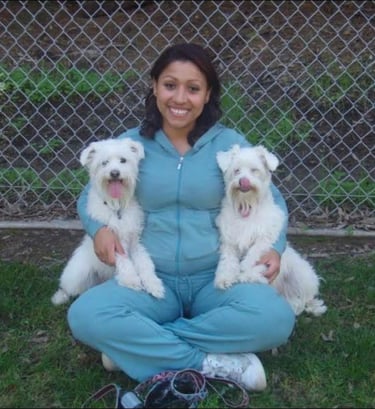 A smiling woman sits cross-legged outdoors holding two fluffy white dogs in front of a chain-link fence.