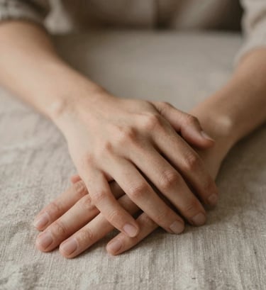 Close-up of hands intertwined softly, resting on a textured linen surface. Warm, natural indoor lighting with earthy tones like #7B6654 and #C8B6A6.