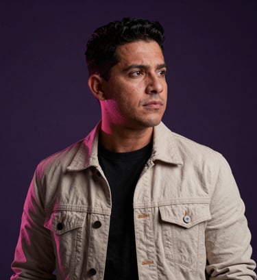 A professional and moody photography portrait of a South American / Venezuelan male musician looking thoughtfully into the distance, wearing contemporary fashion, set against a dark purple studio background with subtle rose-colored rim lighting.