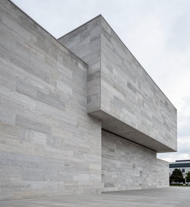 A wide-angle professional shot of a modern museum facade in Northern Europe. Sharp angles of light gray stone against a pale sky. The style is minimalist and high-fashion, emphasizing refined taste and architectural precision.