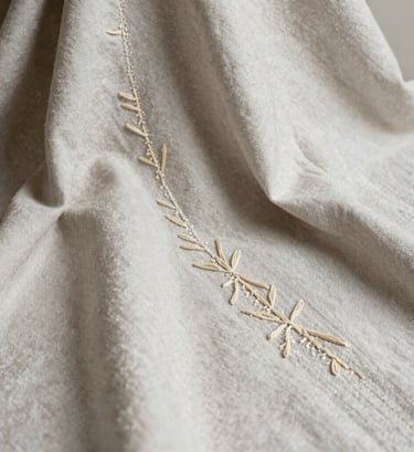 A close-up high-fashion editorial photograph focusing on handmade textile details and natural embroidery on a cream-gray fabric. Soft, diffused lighting highlights the organic textures. Styled in a sophisticated, minimalist South American editorial design layout, emphasizing the raw elegance of natural fibers.