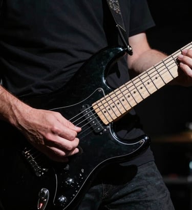 Close-up photography of a musician's hands playing a black electric guitar. Low-key lighting with deep black shadows and off-white highlights on the strings. Professional artistic style, Western European concert setting.