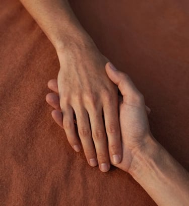 A close-up lifestyle photograph focusing on two hands gently holding each other, with a textured terracotta fabric in the background. Taken in Spain / Southern Europe during the golden hour. The lighting is soft and warm, highlighting the natural skin texture and a sense of deep, intimate connection. Authentic, natural, deeply emotional, cinematic depth of field.