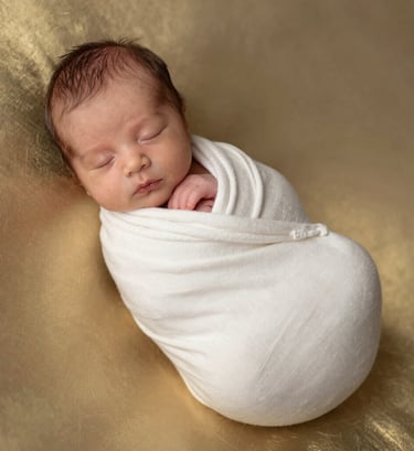A high-end portrait of a sleeping newborn baby wrapped in a soft off-white organic cotton cloth. The baby is resting on a metallic gold-toned surface that reflects soft light. Clean, minimalist composition in a professional Turkish / Istanbul setting.