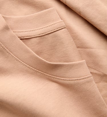 Close-up of a beige crew neck t-shirt showing the ribbed neckline and soft cotton jersey fabric texture.