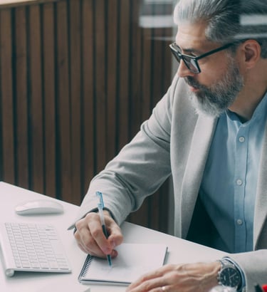 Professional businessman in a grey blazer taking notes while working on a computer in a modern office.