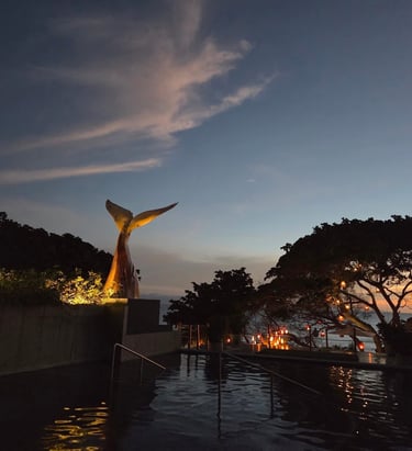 Piscina infinita de lujo al atardecer, con una escultura de cola de ballena y faroles luminosos.