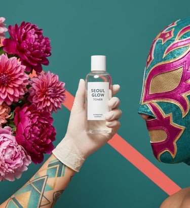 Seoul Glow hydrating toner bottle held by a gloved hand next to pink dahlias and a luchador mask.