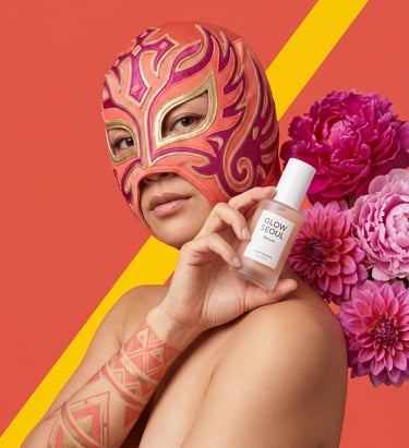 A person in a lucha libre mask holds Glow Seoul facial serum next to pink dahlia flowers.