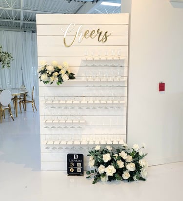 White champagne with white roses and greenery flowers and a sign that says cheers