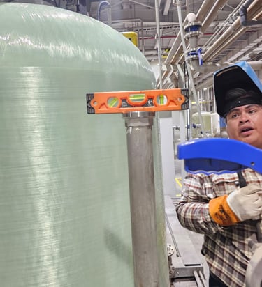 Trabajador con careta de soldar usando un nivel sobre una tubería junto a un gran tanque industrial.
