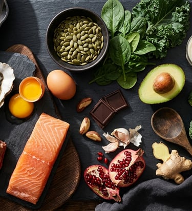 Flat lay of healthy nutrient-dense foods including salmon, steak, avocado, and leafy greens on dark stone.