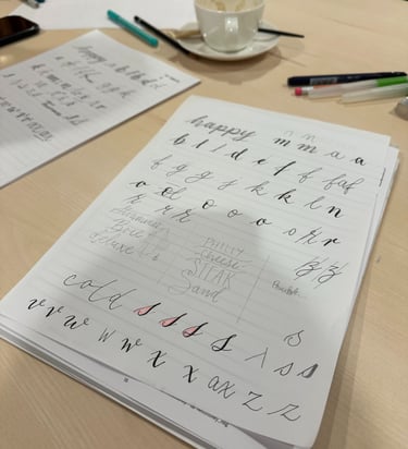 Cursive handwriting and calligraphy practice sheets on a desk with pens and a coffee cup.