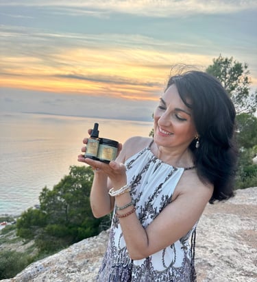 A woman holds natural organic skincare products during a scenic sunset over the Mediterranean ocean.