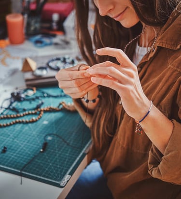 mujer sosteniendo joyeria artesanal frente a mesa de trabajo llena de cuentas e hilo