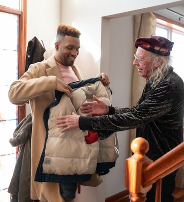 An older man helps a younger man try on a tan winter coat inside a brightly lit home foyer.