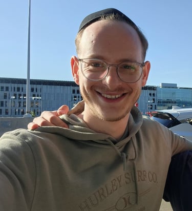 Smiling man with glasses and a yarmulke wearing a tan Hurley hoodie outdoors on a sunny day.