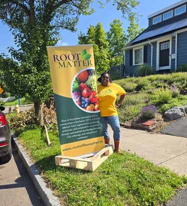 A woman in a yellow shirt stands next to a Root of the Matter banner for urban gardening in Rochester, NY.