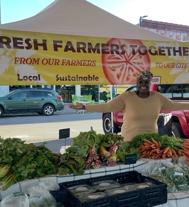 A smiling vendor at a local farmers market stands behind a stall with fresh sustainable vegetables.