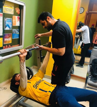 A personal trainer spotting a young man performing a barbell bench press in a gym setting.