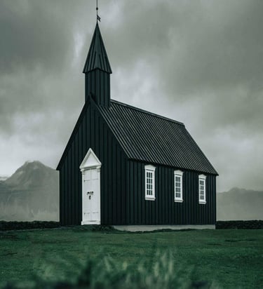 Small black church with white trim and cross-topped steeple set against dramatic cloudy sky.