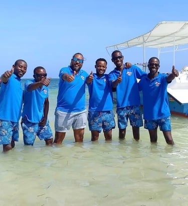 A group of smiling scuba intructors in blue shirts giving thumbs up in the ocean near boats.