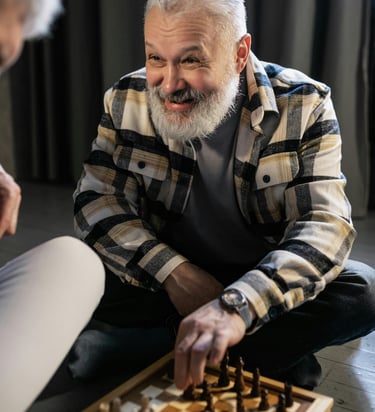 Homem idoso sorridente, com barba branca e camisa xadrez, concentrado em uma partida de xadrez no chão.