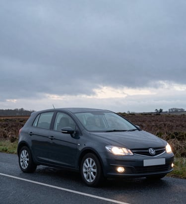 Close documentary frame of a dark saloon car parked on an empty North Lincolnshire road at early morning, headlights off, driver-side door visible, overcast grey sky above flat fenland horizon, natural cold daylight, no people