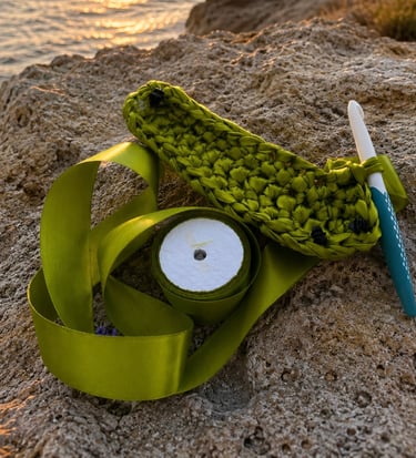 Handmade green crochet ribbon project with a crochet hook resting on seaside rocks at sunset.