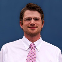 Professional portrait of a young man with glasses and a pink tie against a blue background.