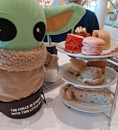 Little Green companion standing beside three tiered plate of mini cakes and tea sandwiches
