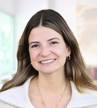 A smiling professional woman with brown hair and gold jewelry wearing a white blazer in an office.