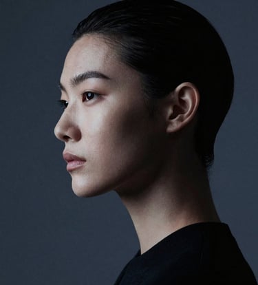 A high-fashion editorial portrait featuring a model in a sharp silhouette. The composition is minimalist, with soft blue-grey lighting hitting the contours of the face against a dark charcoal backdrop. Refined and elegant style.