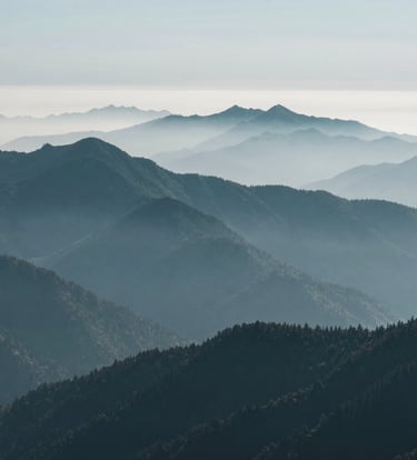 A professional wide-angle landscape photograph of a misty mountain range in the North American Pacific Northwest. The scene is dominated by layers of peaks in dark teal and muted blue-grey, shrouded in soft fog. The composition is minimalist and modern, emphasizing the vast, calm beauty of the wilderness.