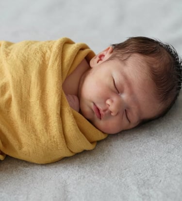 A close-up, high-resolution photo of a sleeping newborn baby wrapped in a soft mustard yellow muslin cloth. The baby is placed on a light grey textured surface. The lighting is soft and natural, suggesting a peaceful Middle Eastern / Turkish nursery setting.