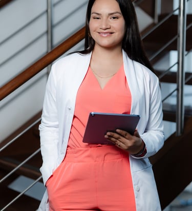 Female healthcare professional in a white lab coat and orange scrubs holding a digital tablet.