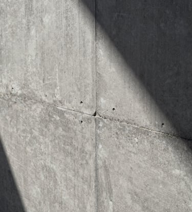 Architectural detail shot of a textured concrete wall with precise joints, showing tectonic rigor. Soft natural light, shadows in dark charcoal grey, material in light grey.