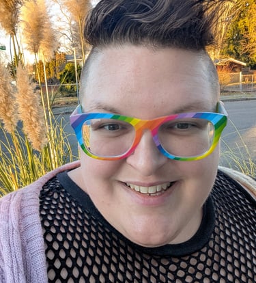 A knitter with short brown hair and rainbow glasses smiling.