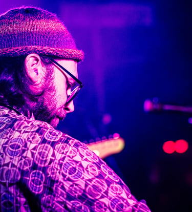 A bearded musician with a beanie plays electric guitar during a live concert with purple stage lighting.