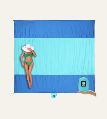 Woman in a green bikini lying on a large sand-free beach blanket with its compact carrying pouch.